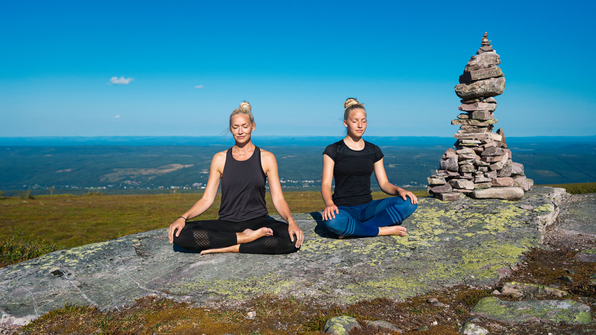 To kvinder udøver yoga i siddende meditationsstilling på en bjergtop i Sverige med en stenhøj i baggrunden og vidstrakt udsigt