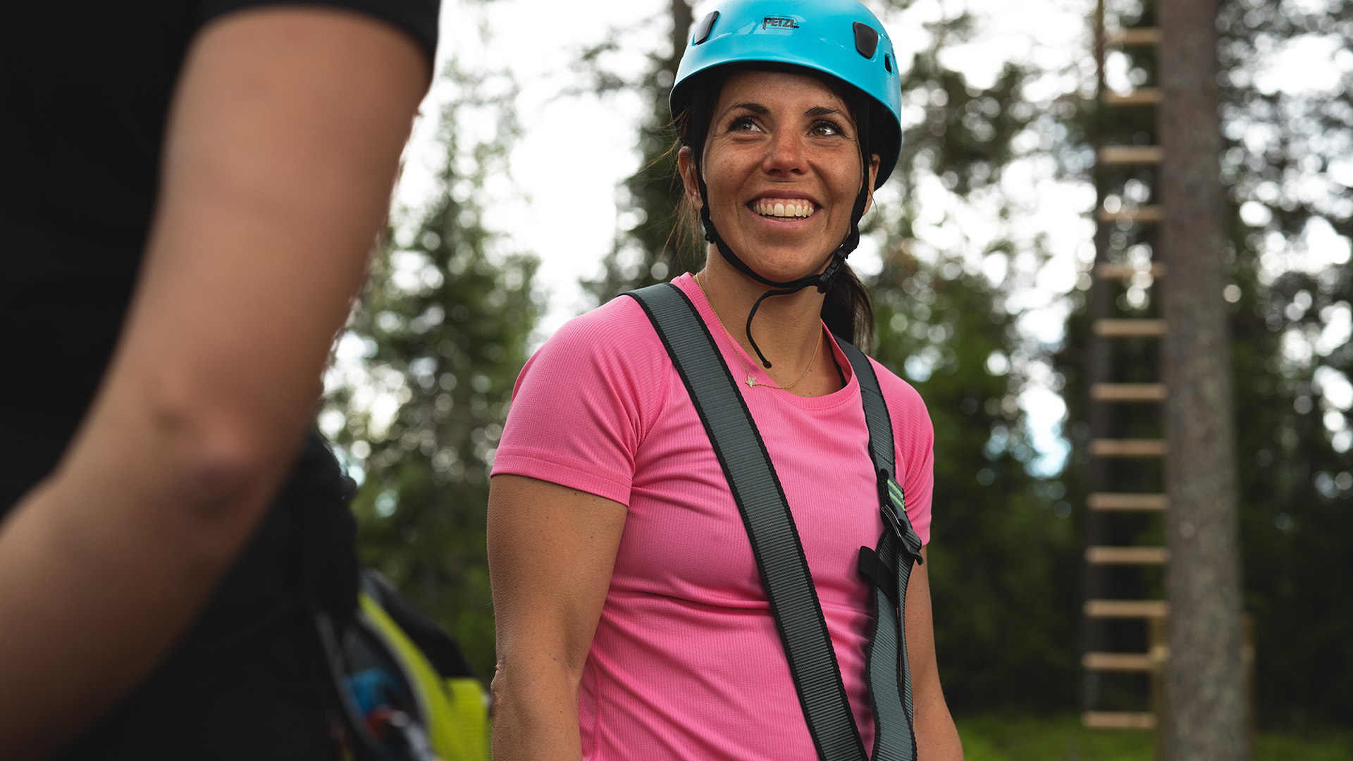 Kvinna i rosa t-shirt och hjälm ler utomhus nära höghöjdsbana i skogsmiljö