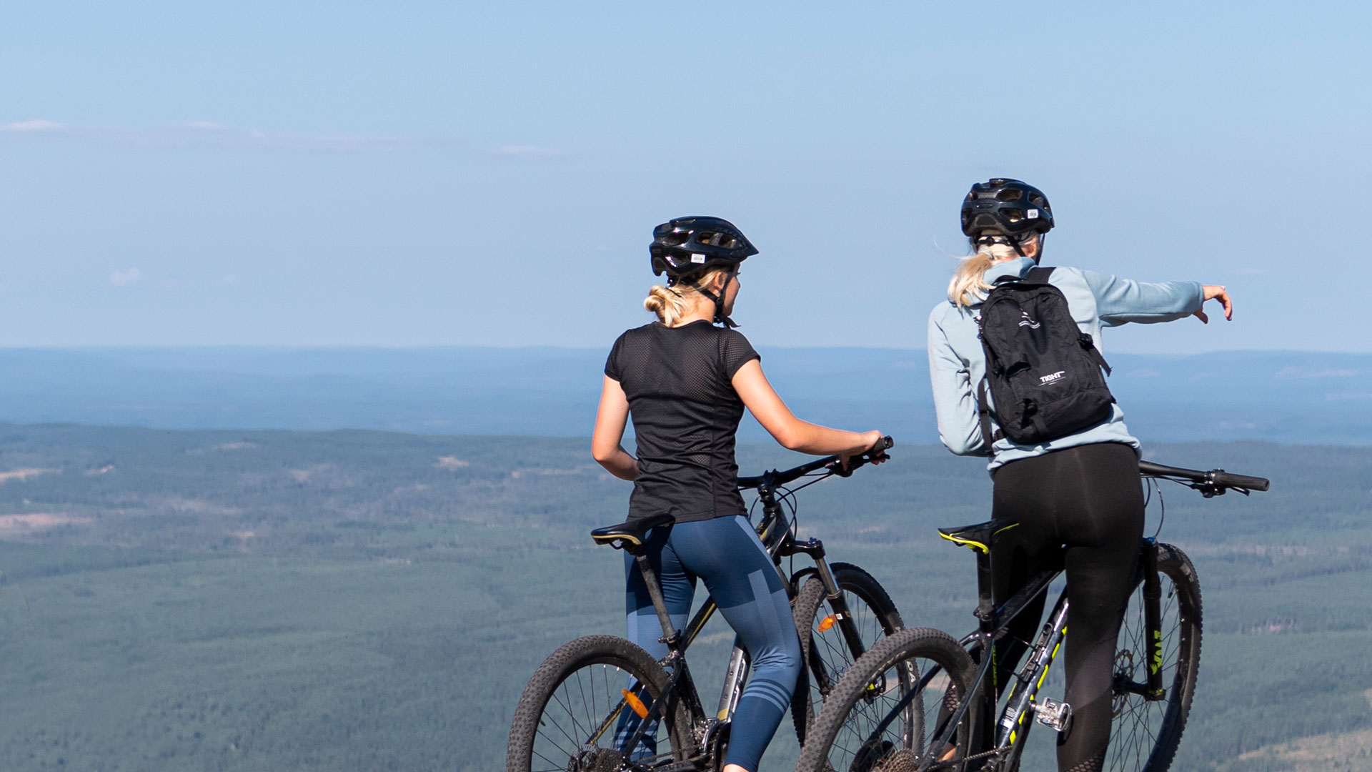 Två personer på mountainbikes beundrar utsikten från en höjd, iförda hjälmar och cykelkläder, med en vidsträckt skog i bakgrunden under en klarblå himmel