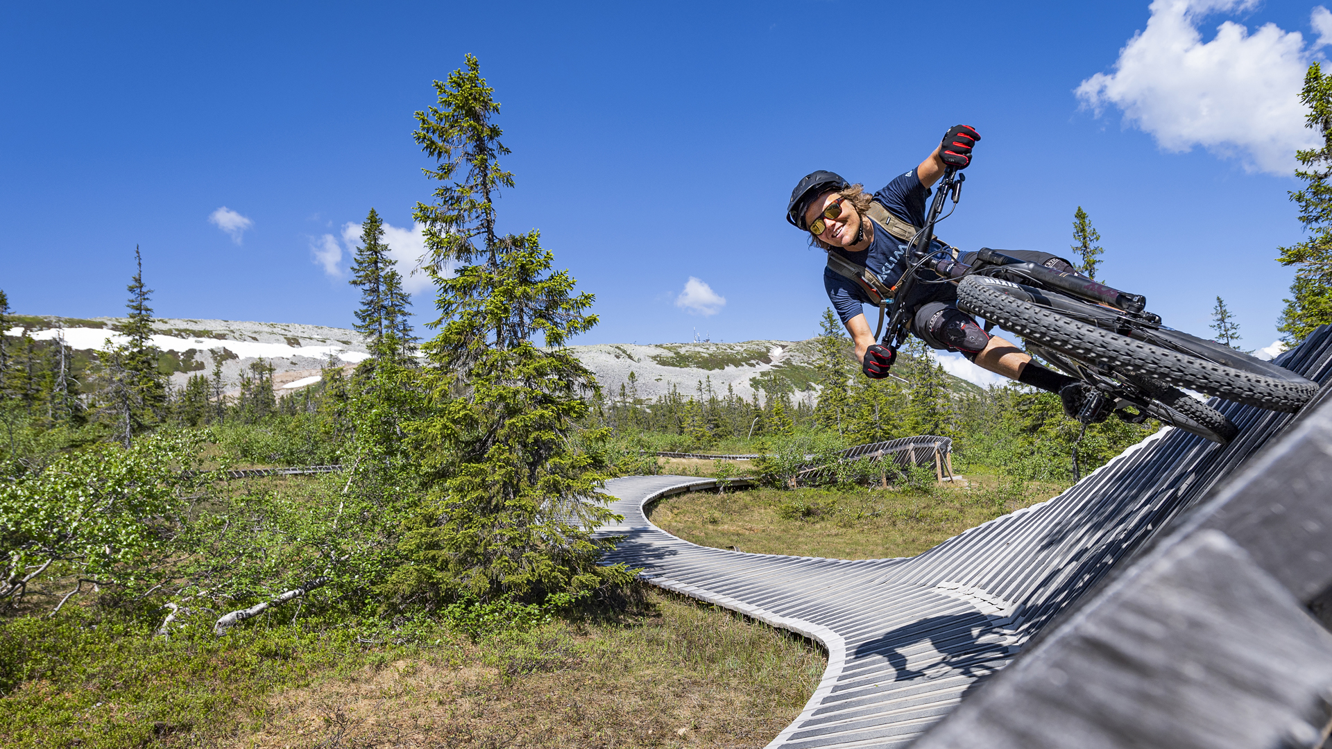 Syklist utfører et triks på en terrengsykkelløype i et skogsområde med noen fjell i bakgrunnen under en klarblå himmel