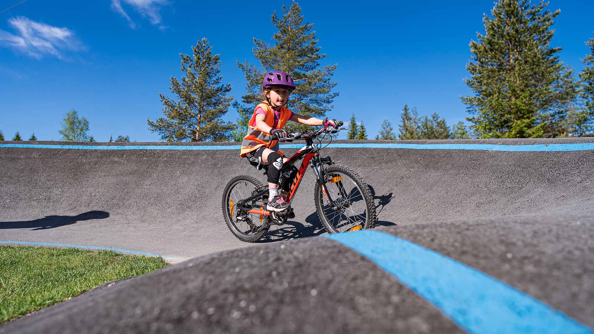 Barn sykler på en pumptrackbane i solskinn, omgitt av trær og blå himmel