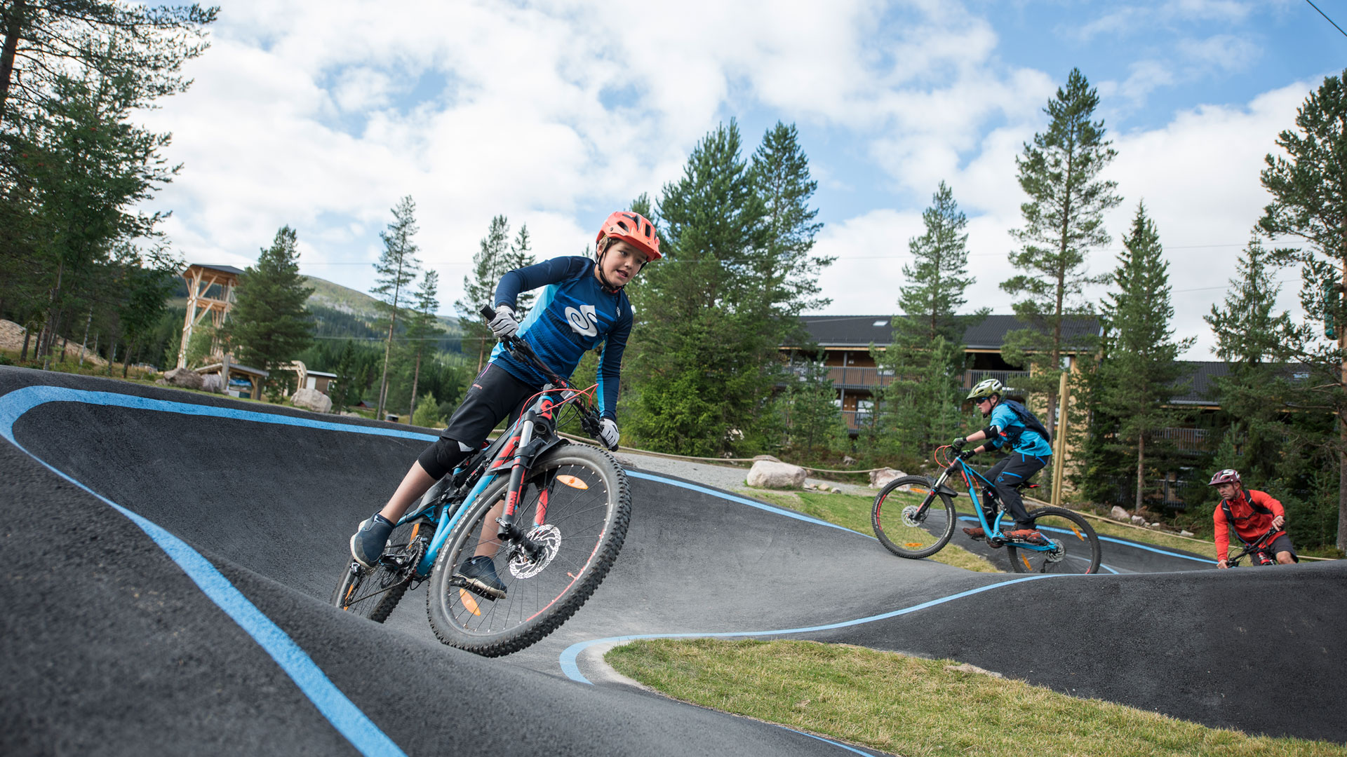 Barn cyklar på en pumptrack i skogen, utrustade med hjälmar och mountainbikes, med träd och en byggnad i bakgrunden