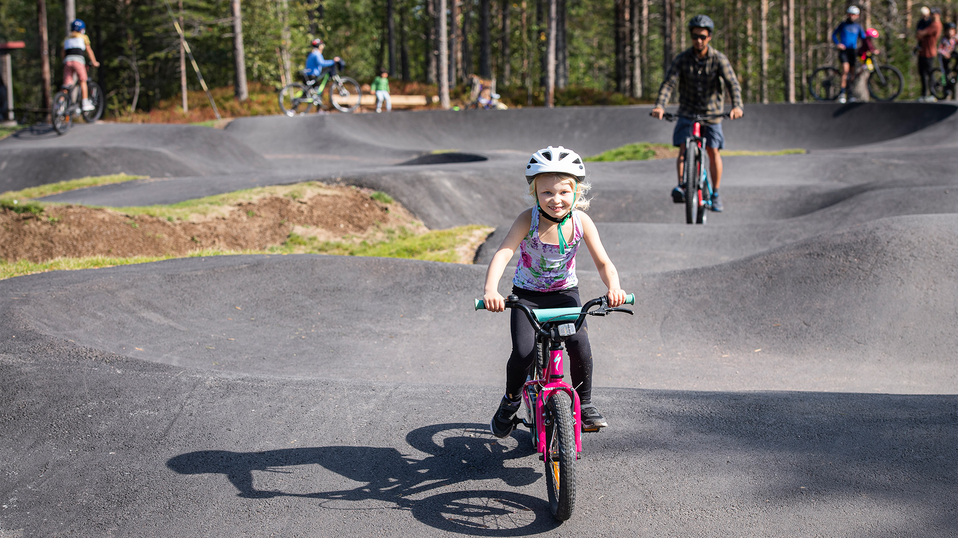 Barn cyklar på pumptrack-bana i skogen, följt av vuxna cyklister