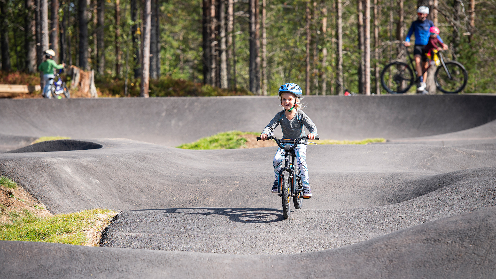 Barn cyklar på pumptrack i skogsmiljö, glatt och lekfullt landskap med andra cyklister i bakgrunden