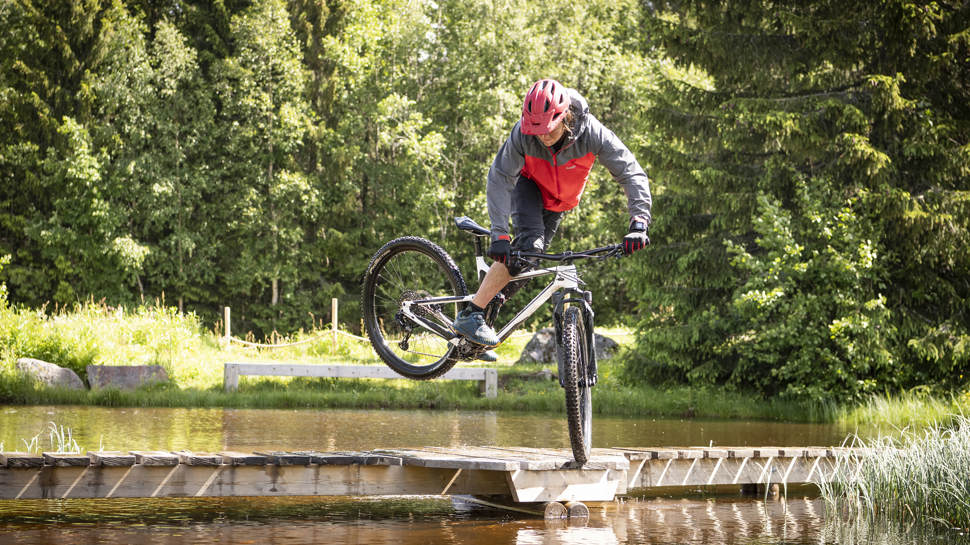 Mountainbikecyklist i röd hjälm och jacka cyklar över en smal träbro i skogen och gör ett tricks