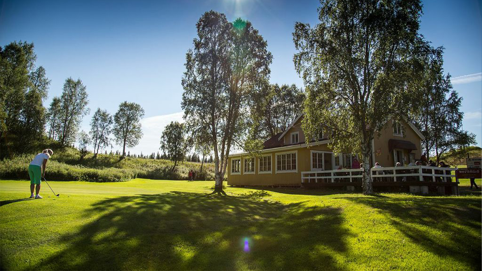 Golfer on a green course near a yellow building surrounded by trees under a clear blue sky