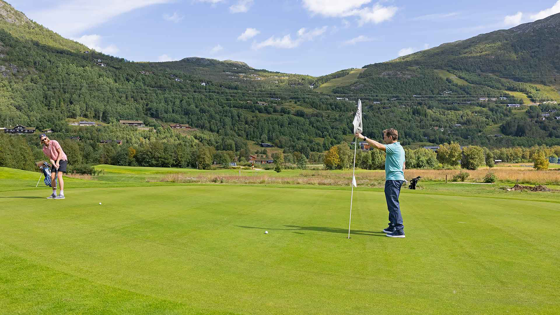 Personer spelar golf på en grön bana med bergig bakgrund under en klarblå himmel