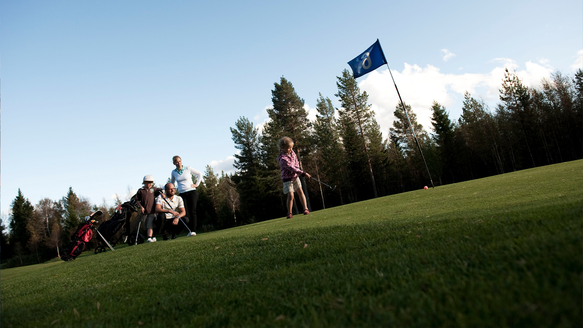 Personer spelar golf på en grön bana omgiven av skog, med en blå flagga synlig i bakgrunden under en klar himmel