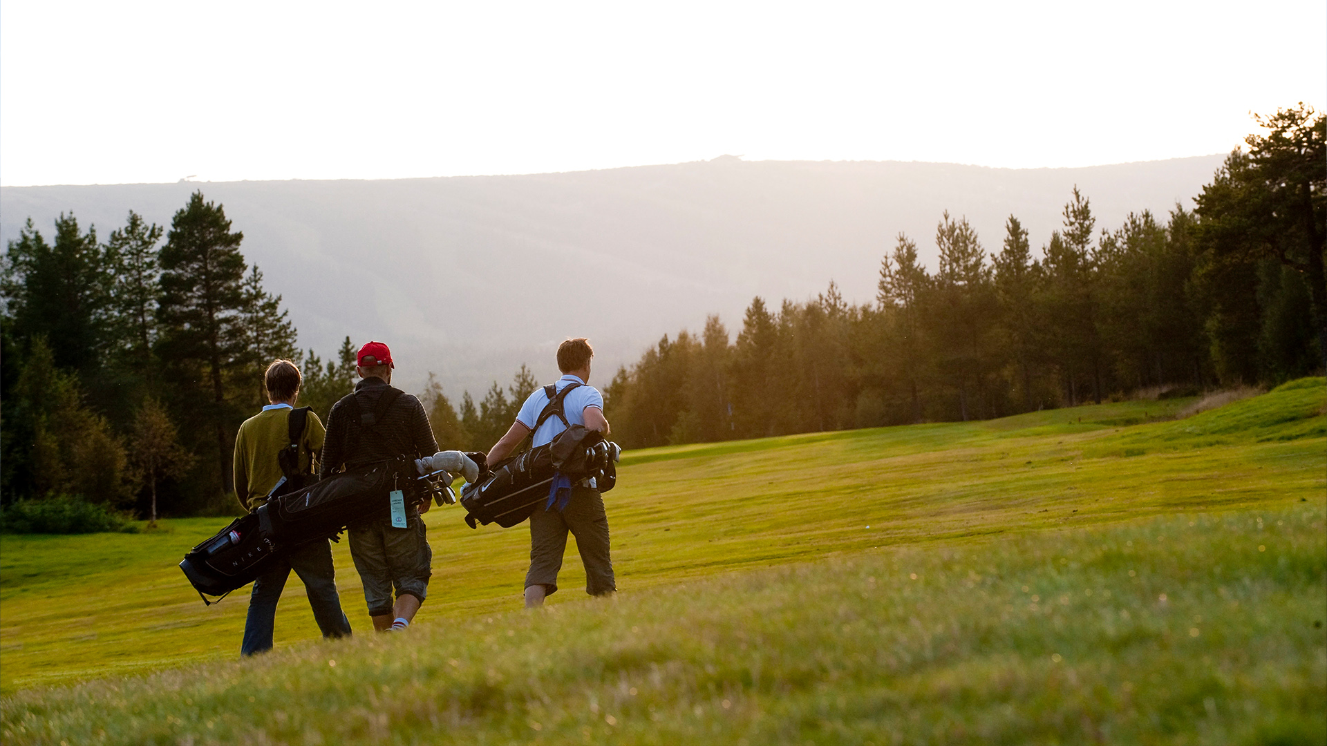 Tre personer går på en golfbana i solnedgången, med skog och berg i bakgrunden