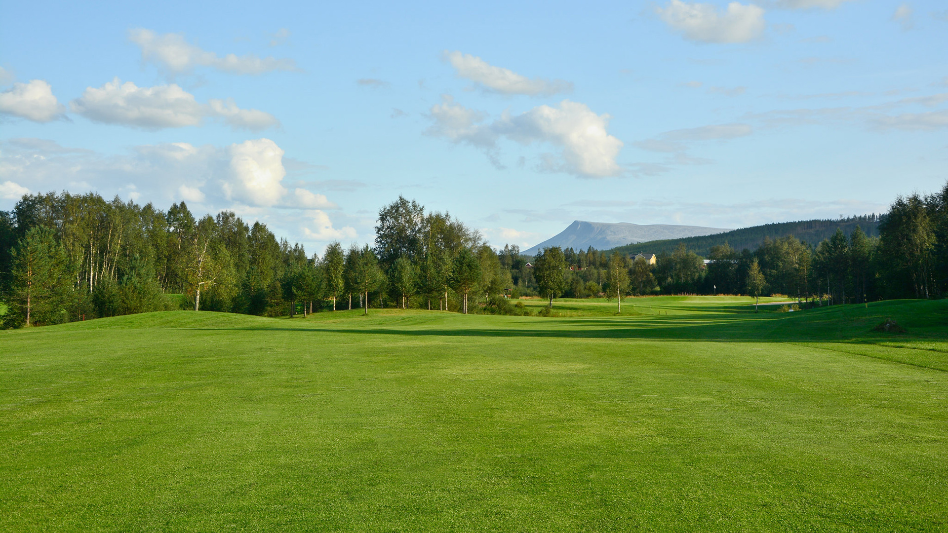 Golfplatz mit grünem Gras, umgeben von Bäumen und Bergen im Hintergrund unter klarem blauen Himmel