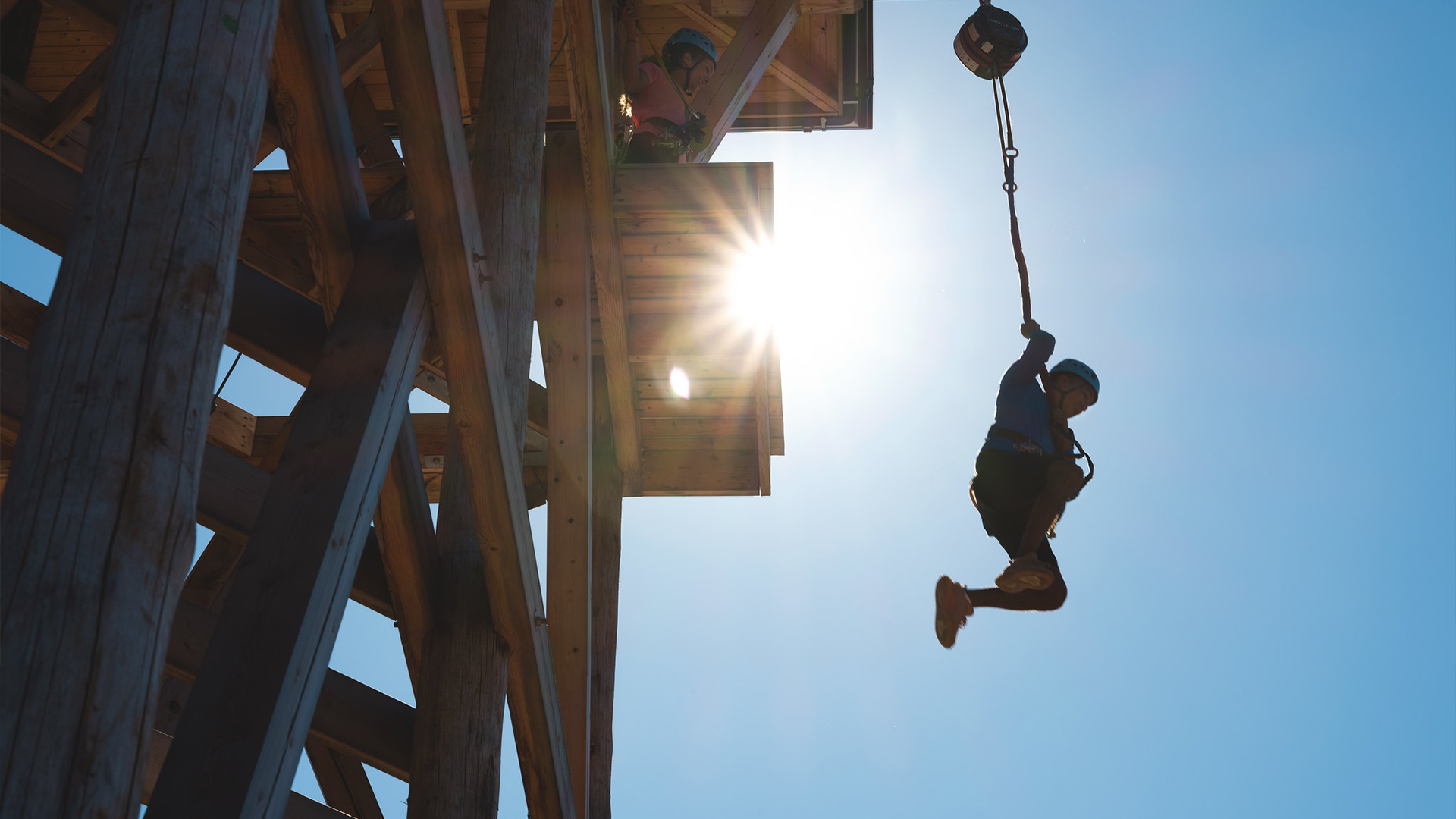 A person rides a zip line from a high wooden platform with the sun backlighting the scene