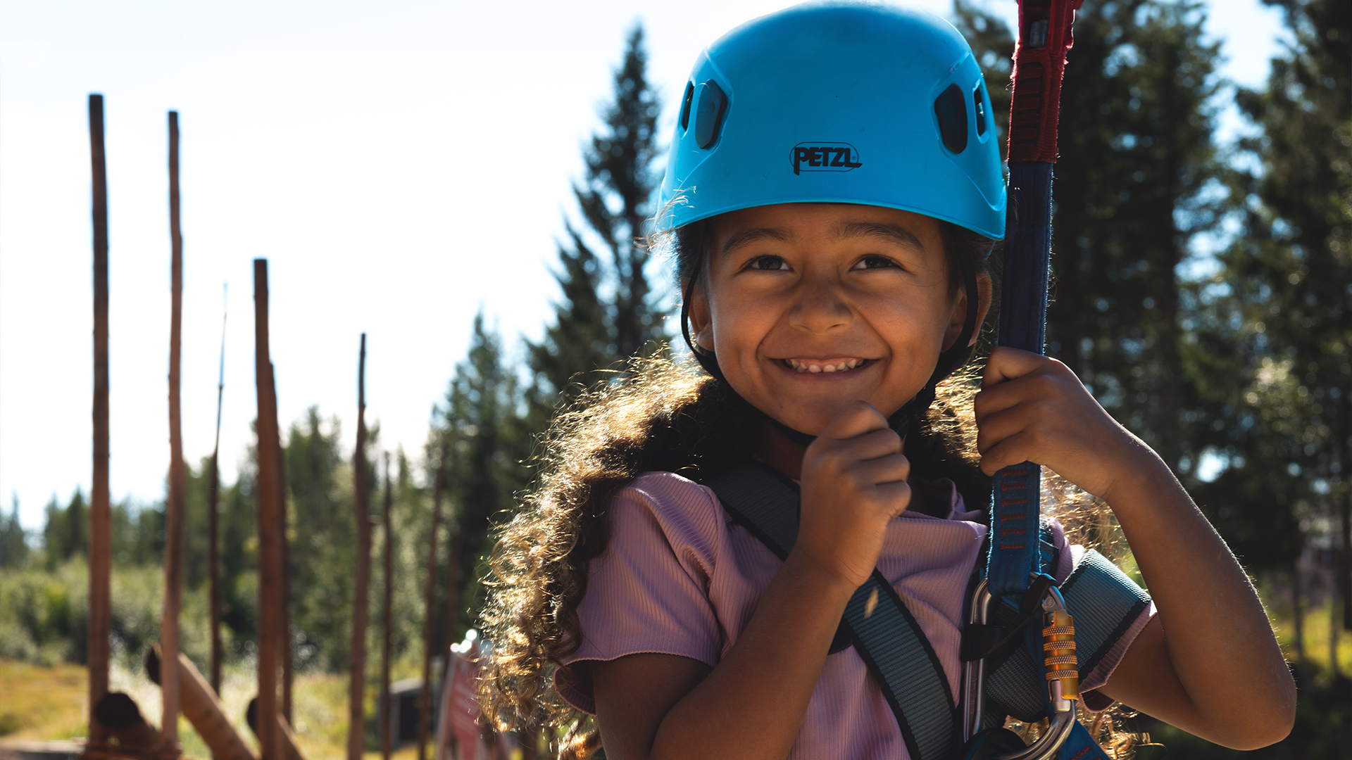 A smiling girl in a climbing harness and blue helmet outdoors in a forest setting