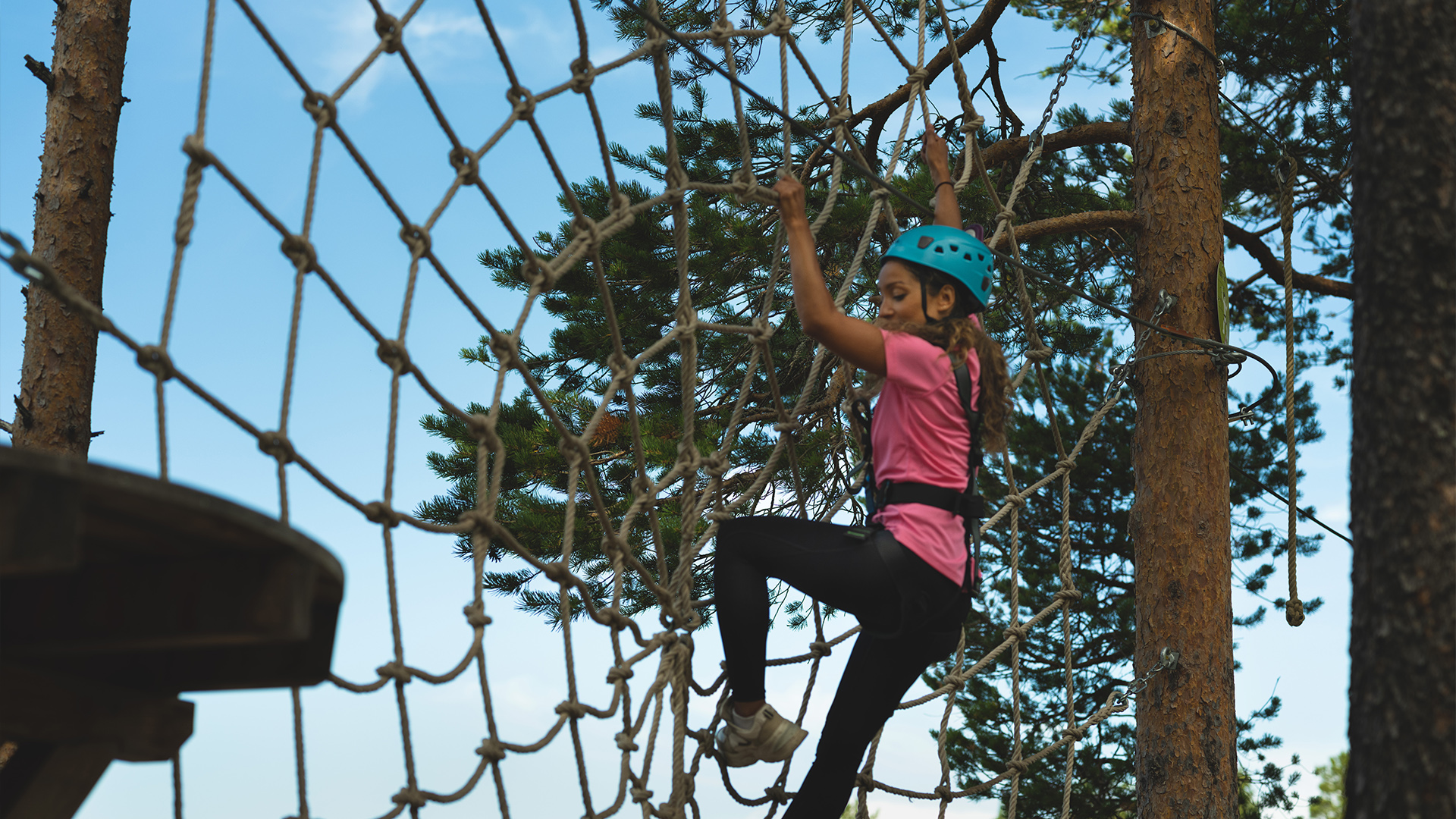 Children climb on a rope net in a high ropes course, wearing helmets and safety harnesses, with trees and a blue sky in the background