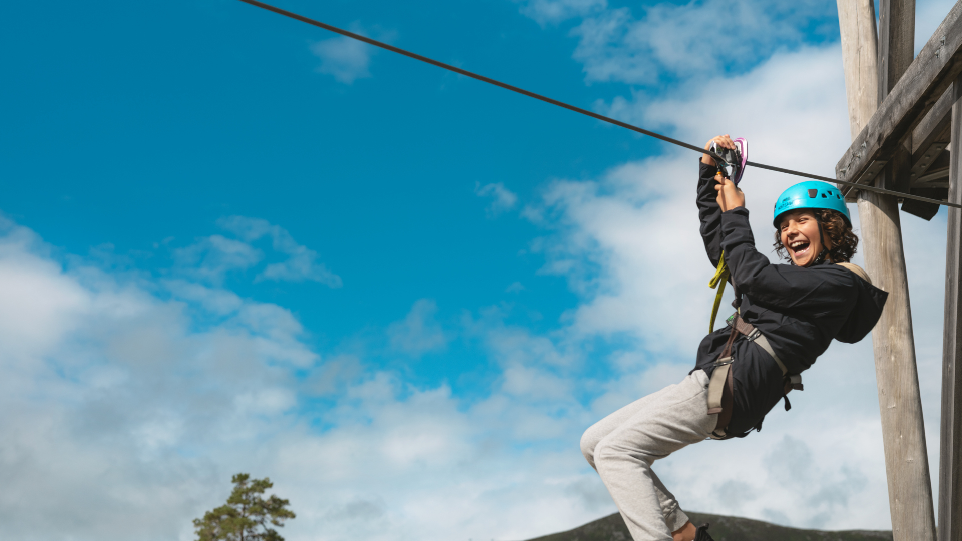 Person på zipline iført blå hjelm, smilende og klar til eventyr under blå himmel