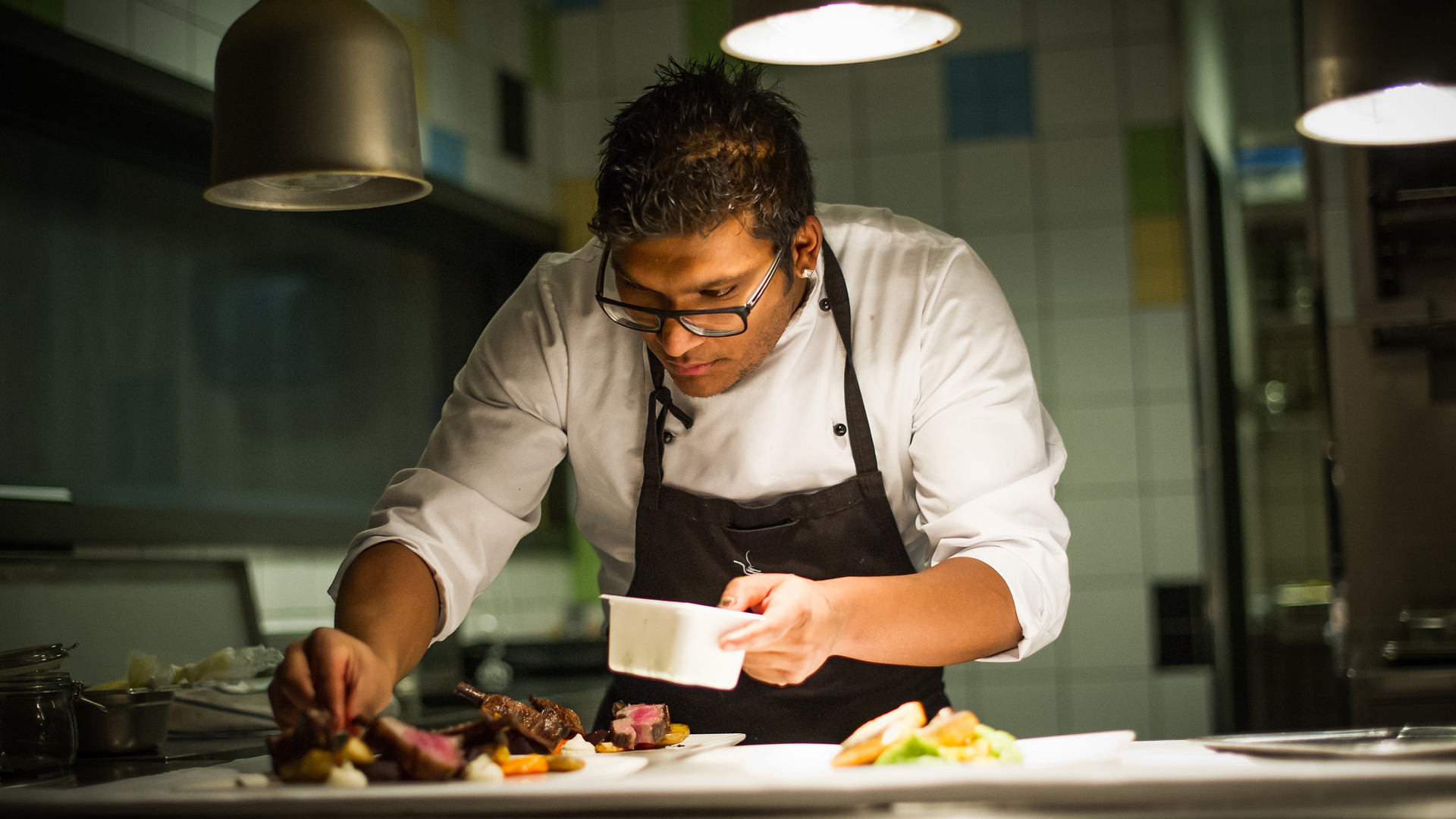 Kok iført hvid kokkejakke og sort forklæde tilbereder en ret i et professionelt restaurationskøkken