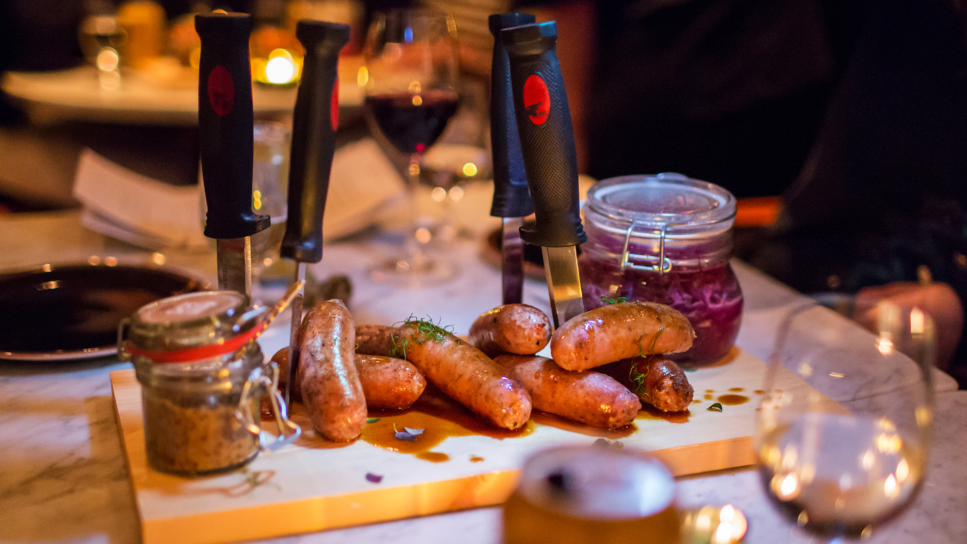 En træbakke med grillede pølser, serveret med rødkål og grov sennep i en krukke, dekoreret med knive på et restaurantbord