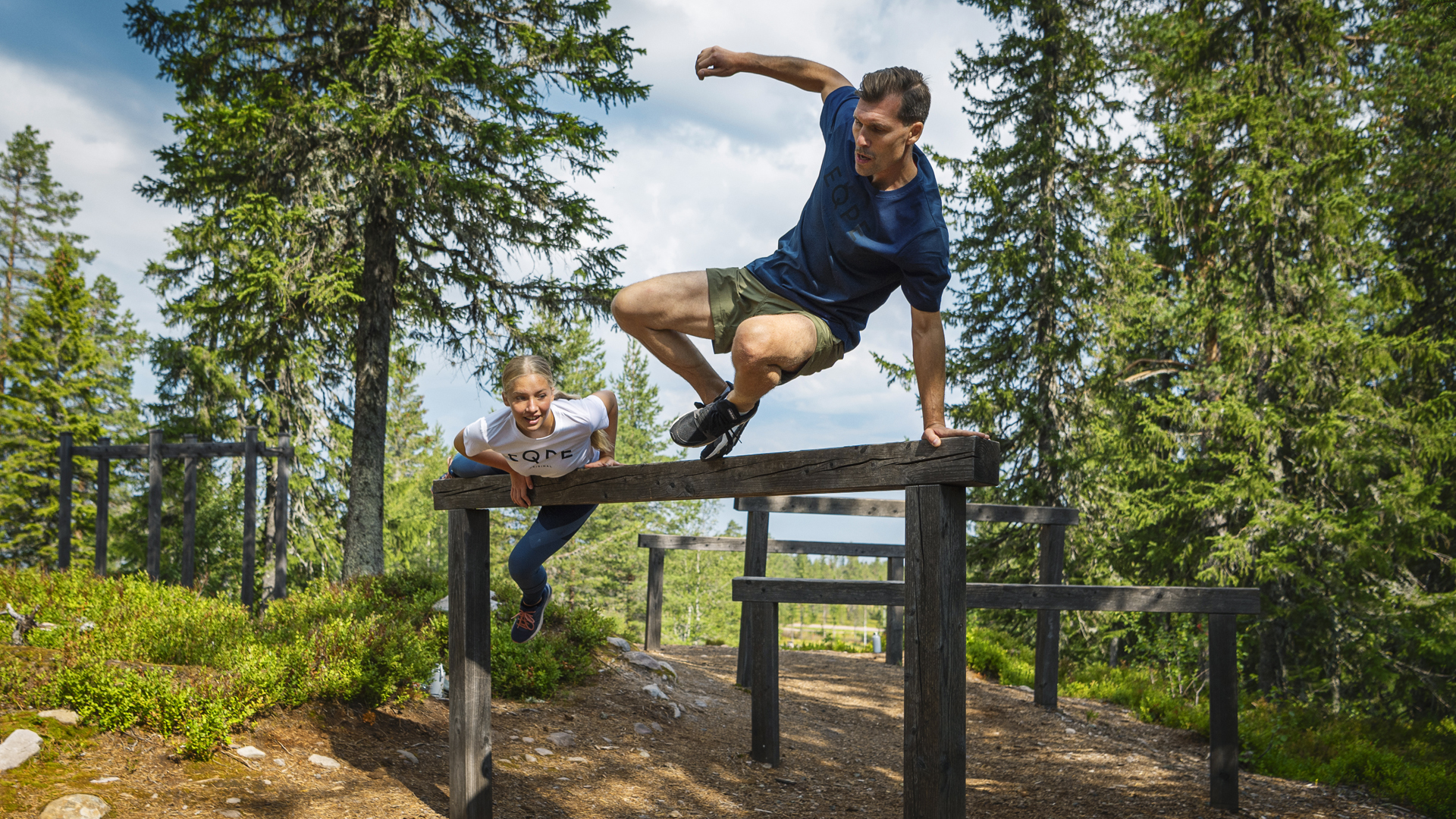 Far og søn hopper over forhindringer på en udendørs forhindringsbane i skoven, en solrig dag med frodige træer i baggrunden