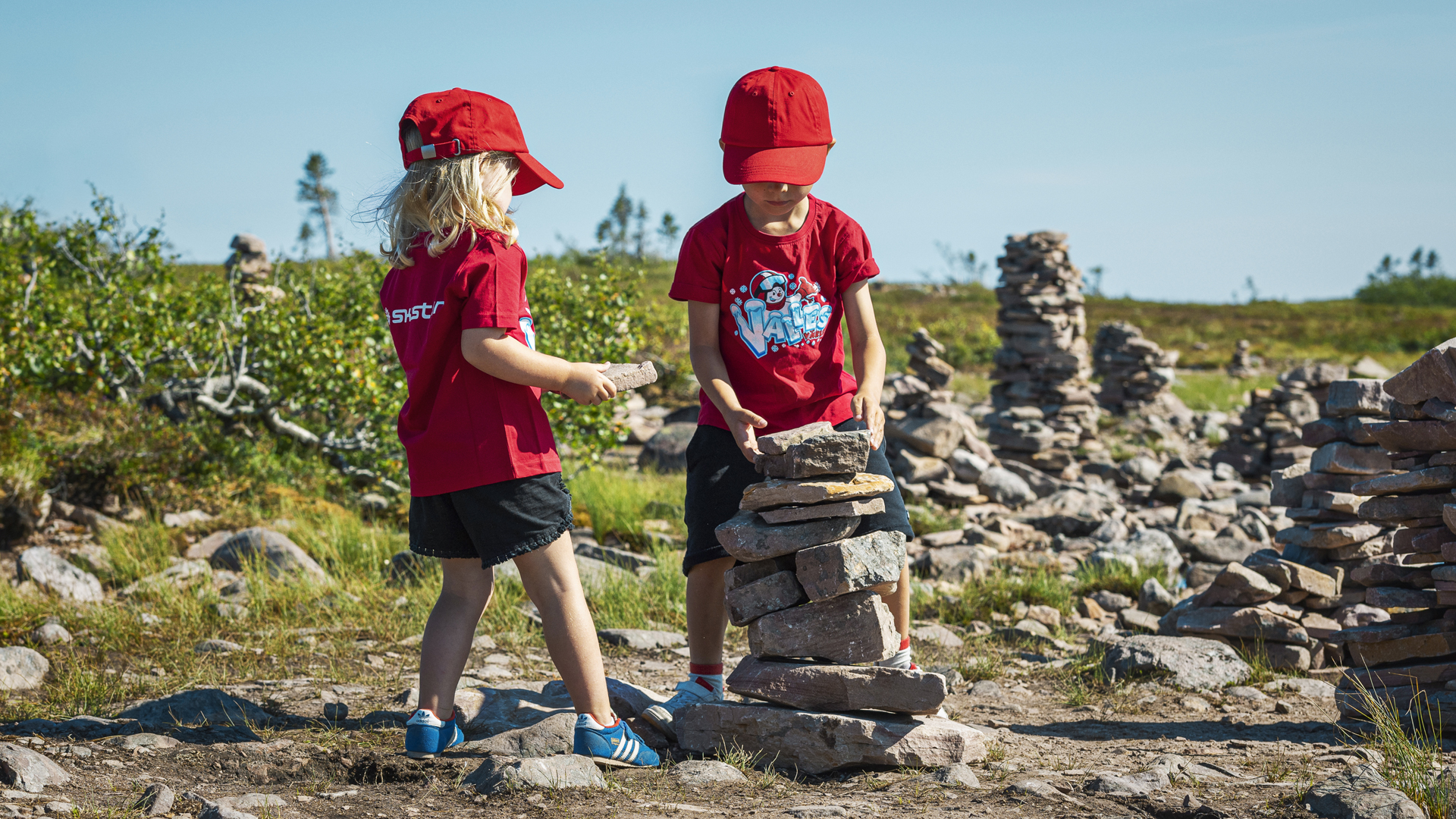 Två barn i röda kepsar och tröjor bygger en stenhög i ett naturlandskap med stenformationer