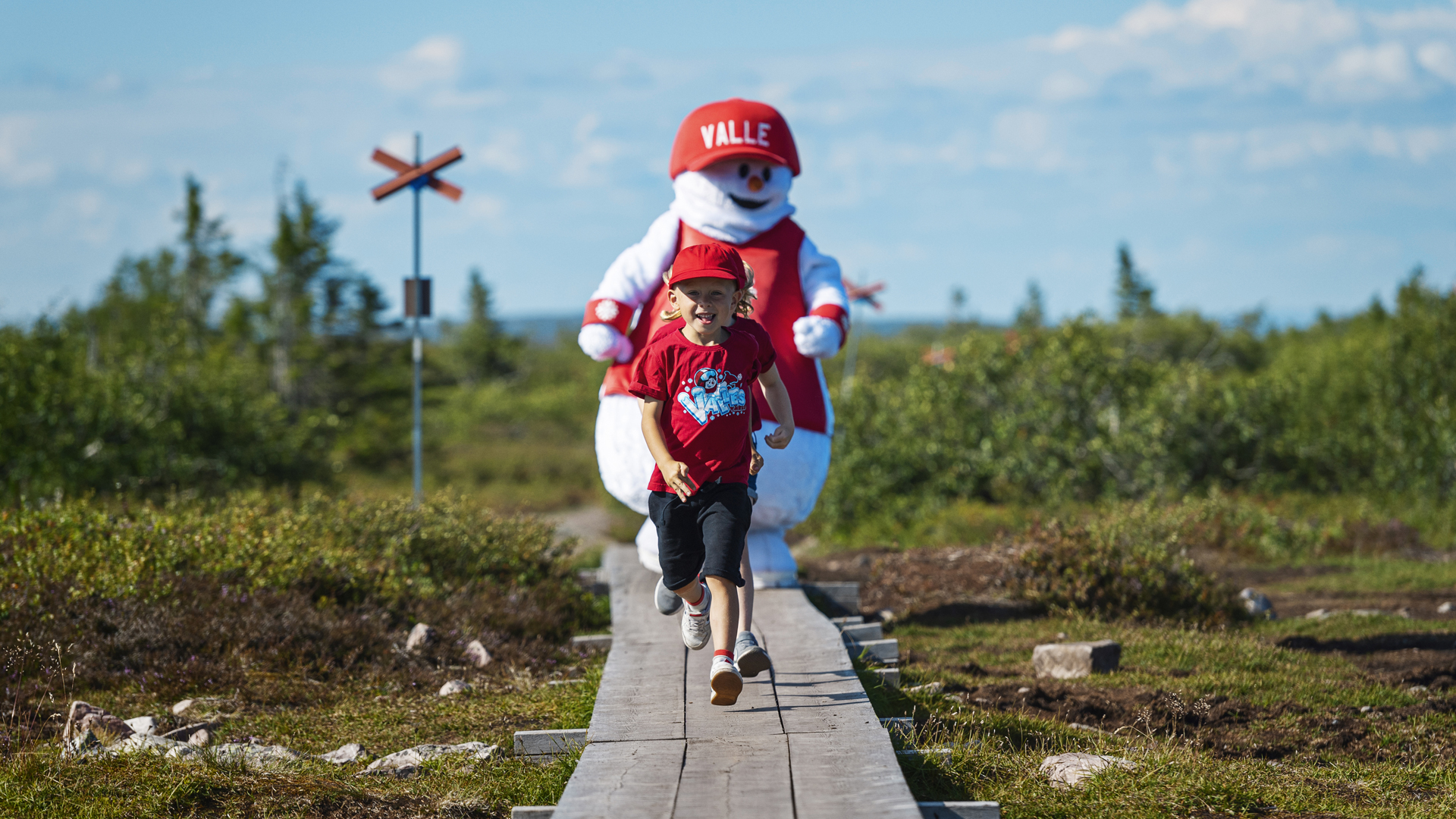Barn springer på en trägångväg i fjällen, följt av en snögubbe i röd keps på en solig dag