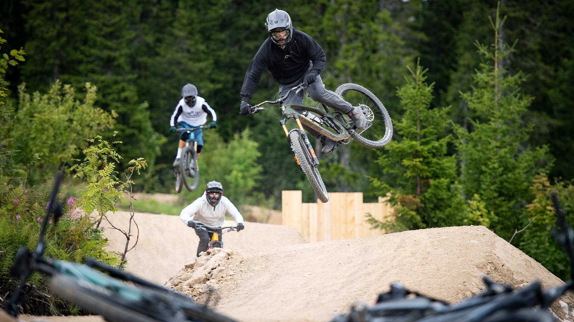 Cyklist utför trick i luften på mountainbikebana, omgiven av skog. Två cyklister följer efter i bakgrunden, sommaräventyr i naturen