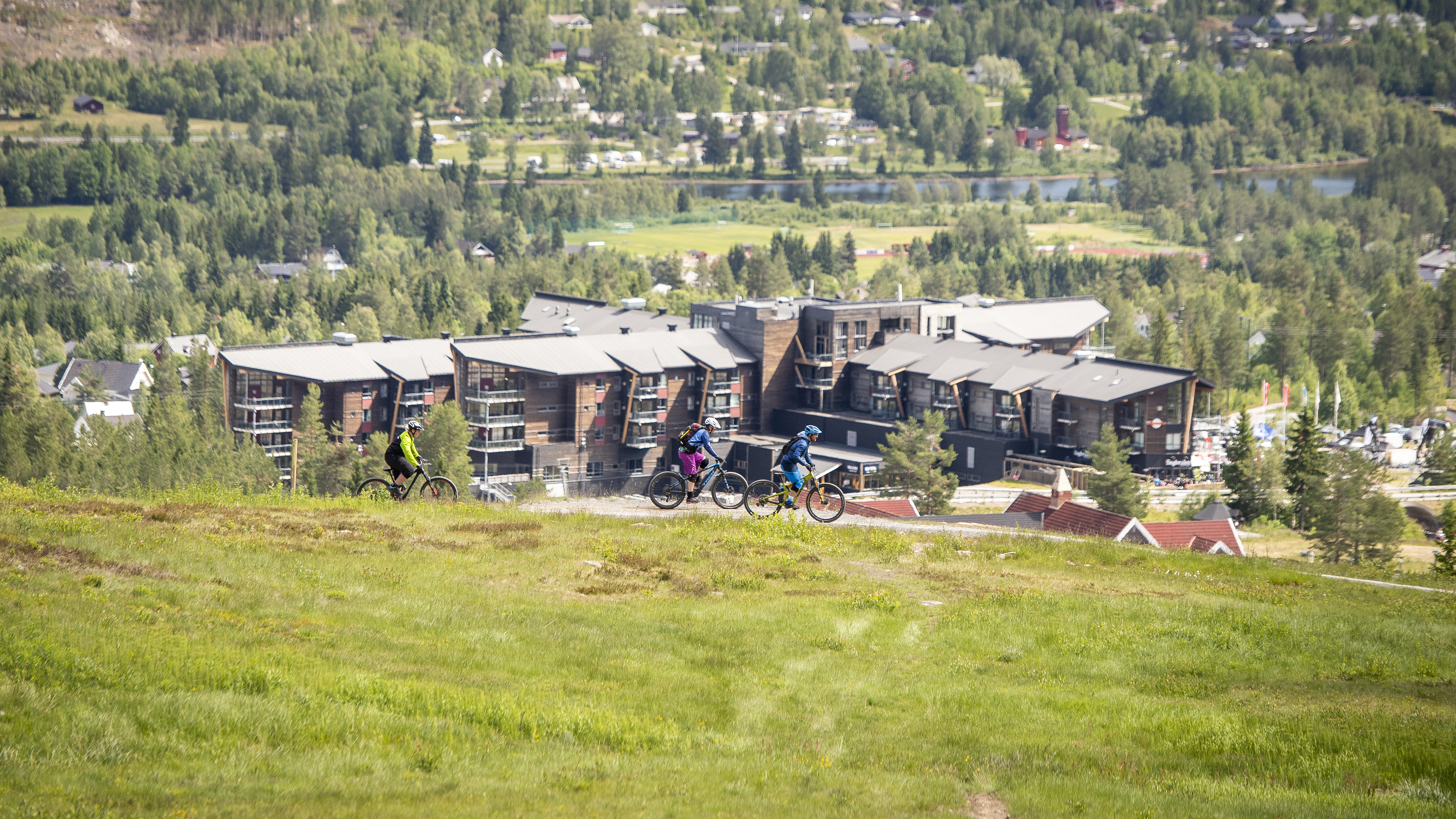 Tre syklister på stisykkel i grønt landskap ved en moderne bygning med skog og fjell i bakgrunnen, sommerdag i Norge