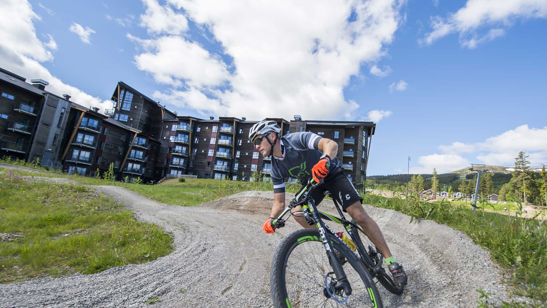 Person cyklar på en grusväg framför moderna lägenheter i naturskön miljö, soligt väder
