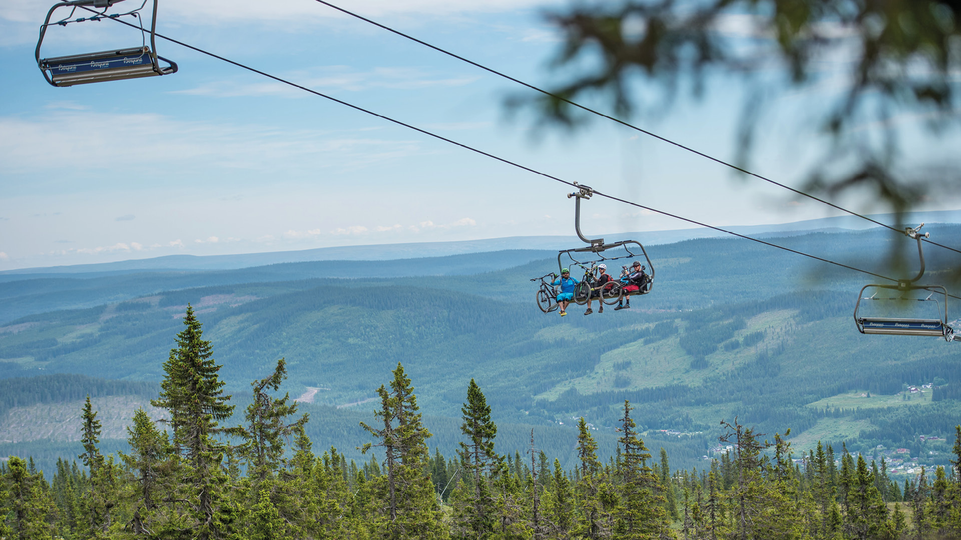 Syklister på stolheis over skogkledd fjellområde i sommerlandskap, med utsikt over dalen