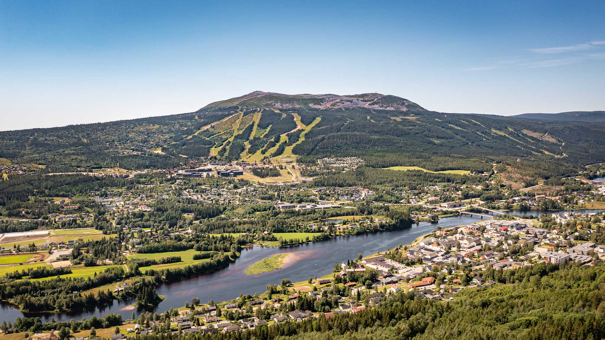 Panoramautsikt over Innbydga, Trysil med fjell i bakgrunnen, grønne skoger og en svingete elv under en klarblå himmel