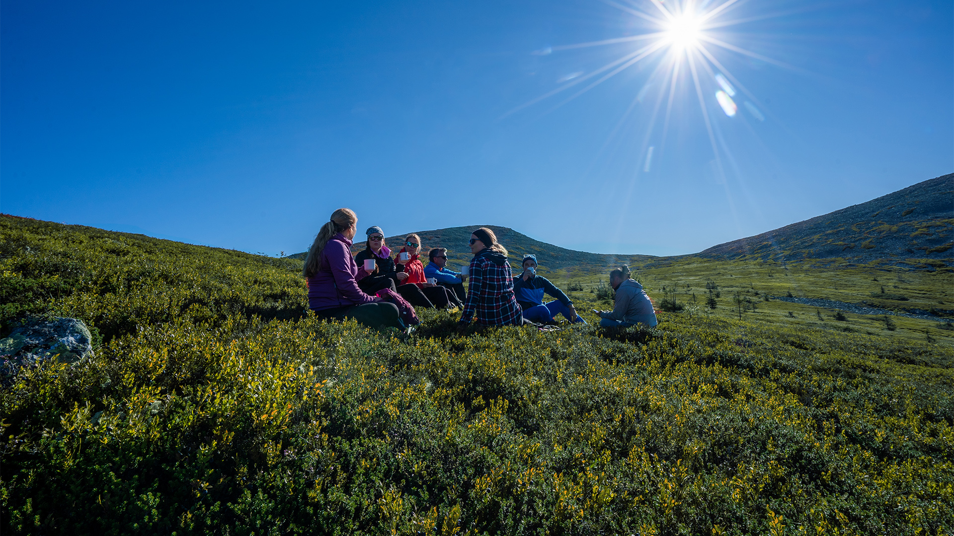 En grupp människor sitter på en grön kulle under klarblå himmel med starkt solsken i fjällmiljö