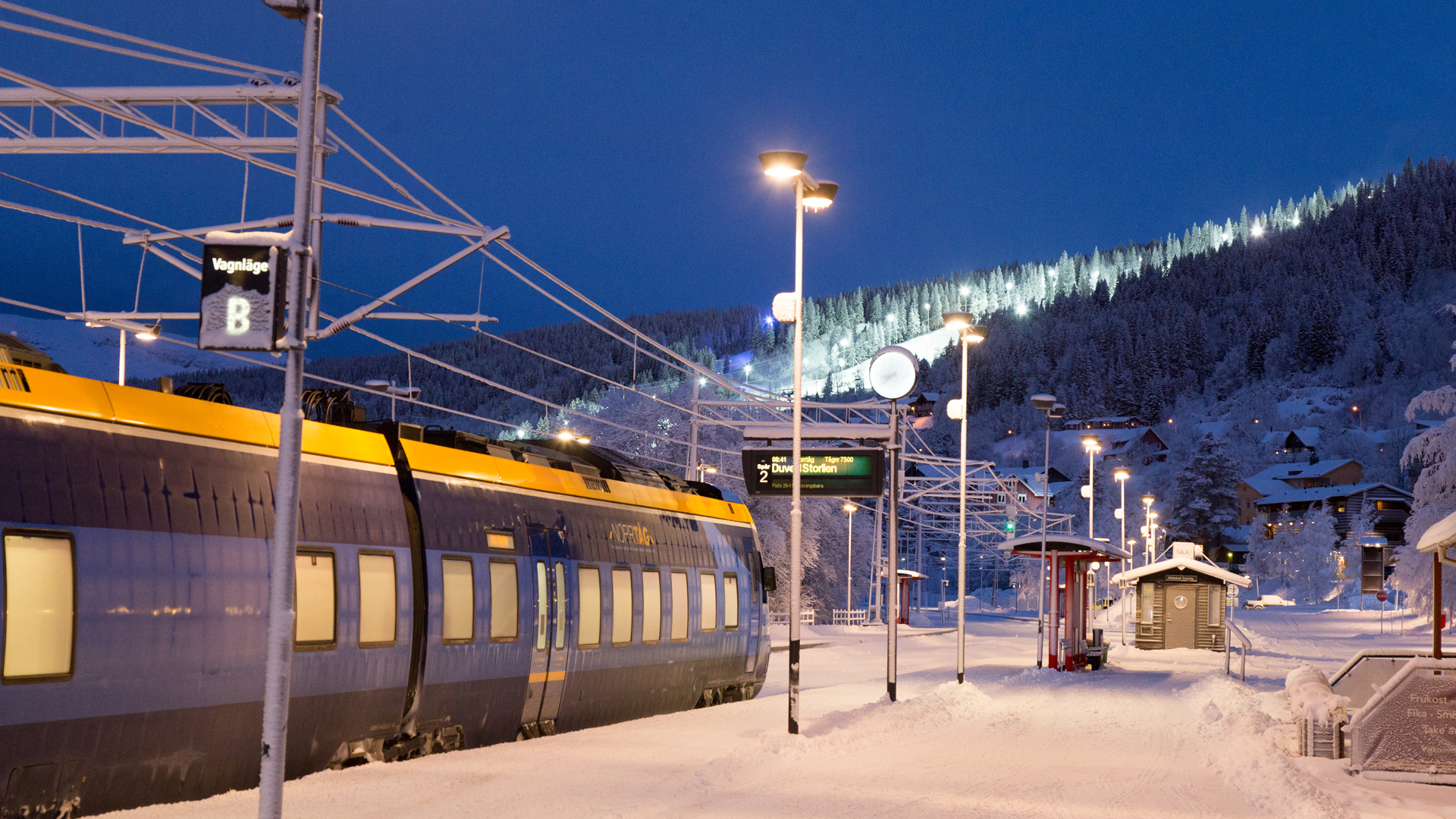 Tog ved en snedækket jernbanestation om vinteren i Sverige, med oplyste skipister i baggrunden og vinterlandskab