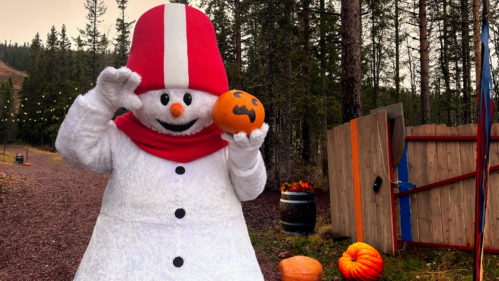 Snögubbe med röd hatt och halsduk håller en pumpa med målade ansiktsdrag i en skogsmiljö under hösten