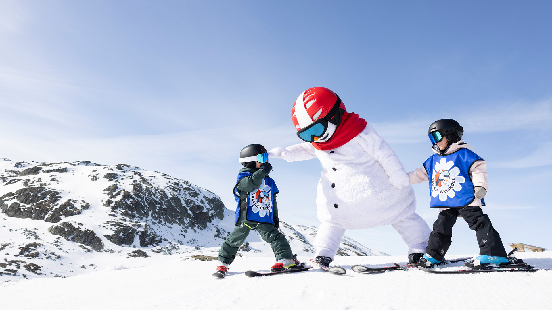 Barn på skidor med en snögubbe-maskot på fjället, under en solig vinterdag
