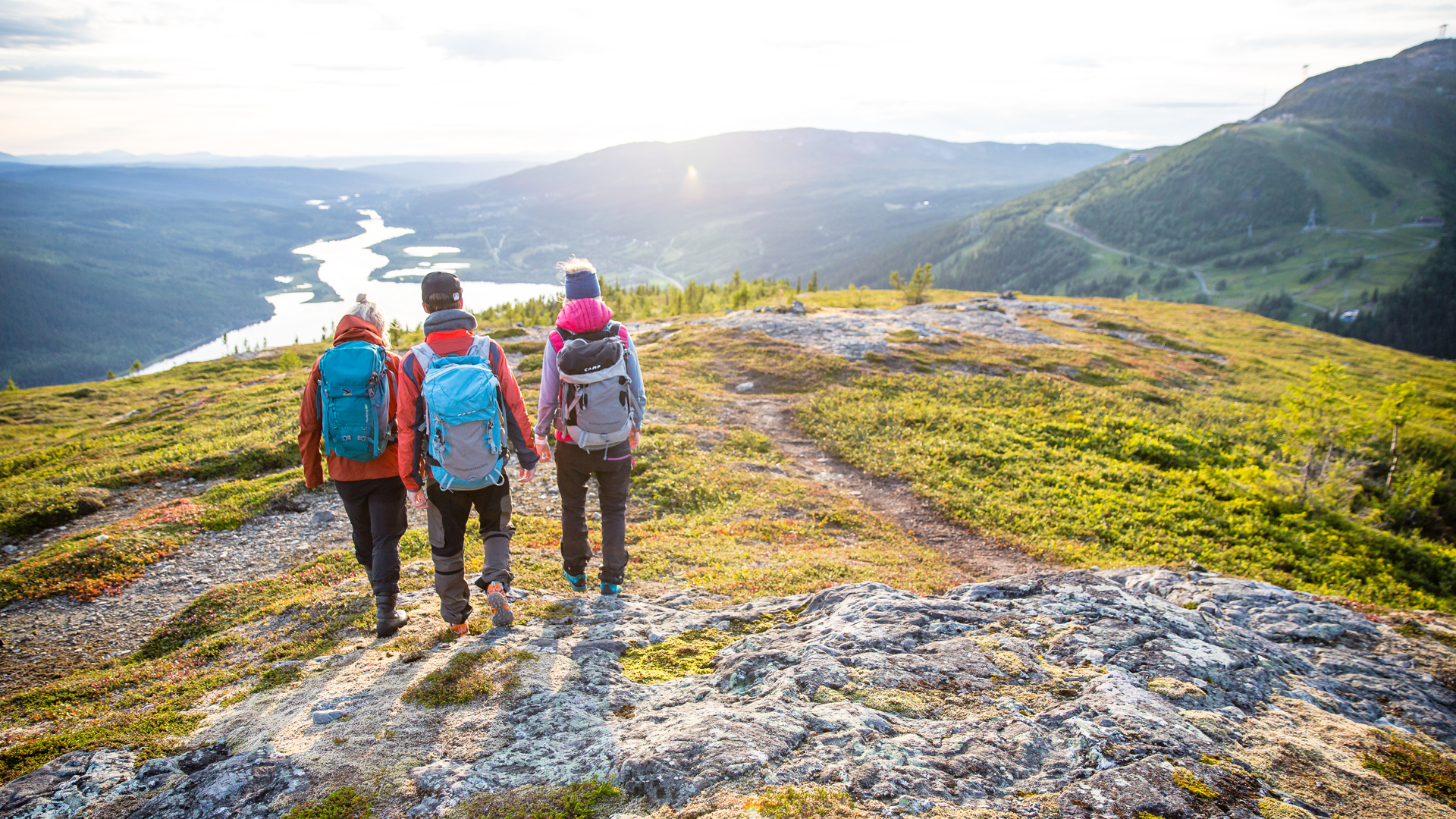Tre vandrare med ryggsäckar på en bergstig med utsikt över en flod och skogsbeklädda dalar i solnedgången, Åreskutan, Sverige