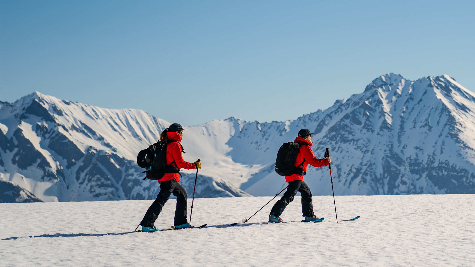 To skiløpere i rød jakke vandrer på en snødekket fjelltopp med majestetiske snøkledde fjell i bakgrunnen på en klar dag