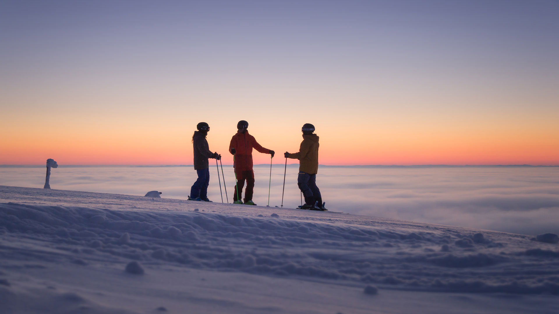 Tre personer i skiantrekk står på en snødekt fjelltopp ved solnedgang, med skylaget under og himmelen i pastellfarger